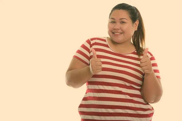 Студийный снимок молодой счастливой толстой азиатки, улыбающейся и показывающей большие пальцы вверх — стоковое фото