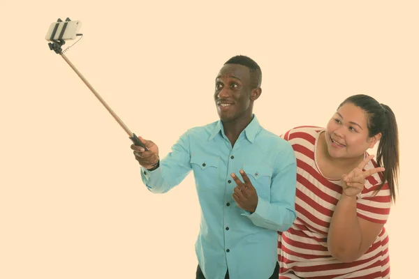 Happy young black African man and young fat Asian woman smiling while giving peace sign and taking selfie picture with mobile phone on selfie stick together