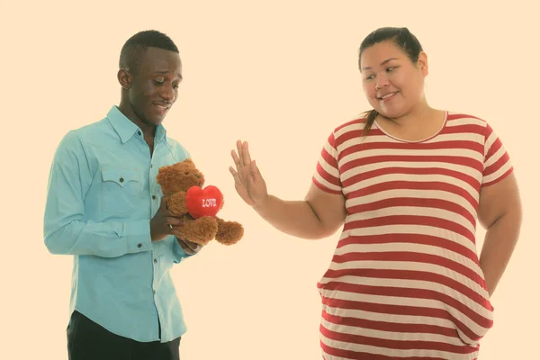 Happy young black African man giving teddy bear with heart and love sign to young fat Asian woman smiling and showing stop sign — Stock Photo, Image