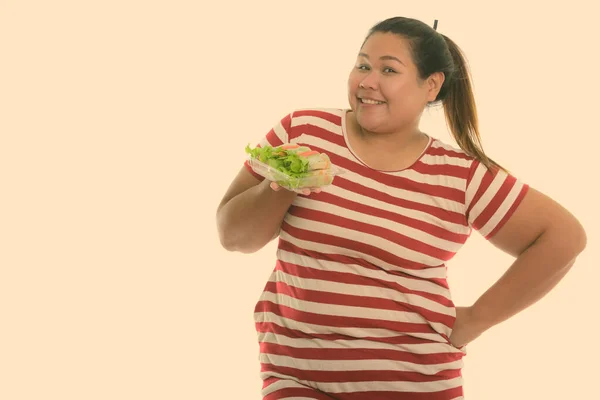 Студийный снимок молодой счастливой толстой азиатской женщины, улыбающейся и позирующей, держа в руках свежую овощную обертку — стоковое фото