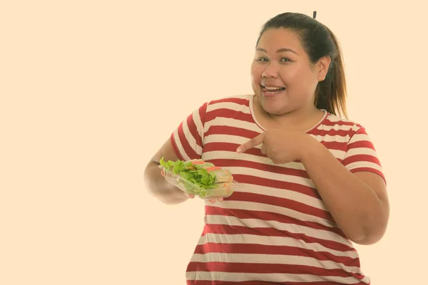 Студийный снимок молодой счастливой толстой азиатской женщины, улыбающейся, держа в руках и указывая на свежую овощную обертку, высунув язык — стоковое фото