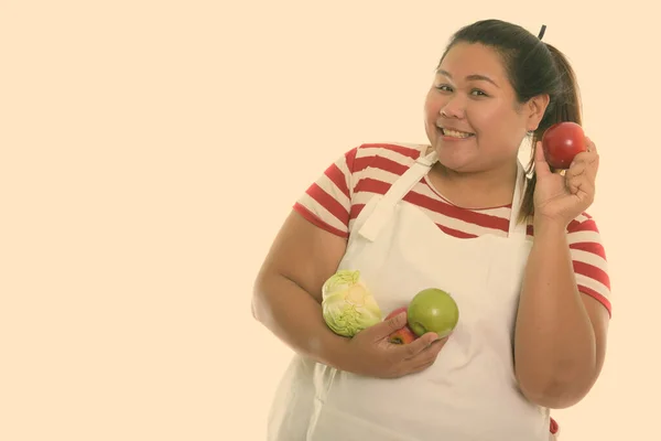 Studio colpo di giovane donna asiatica grassa felice sorridente mentre tiene mela rossa con frutta e verdura mentre indossa grembiule — Foto Stock