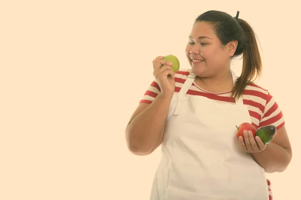 Studio colpo di giovane felice donna grassa asiatica sorridente e guardando mela verde, mentre tenendo frutta fresca con grembiule — Foto Stock