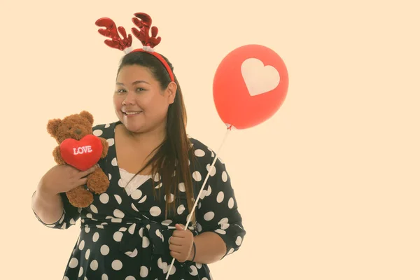 Студийный снимок молодой счастливой толстой азиатской женщины, улыбающейся, держа плюшевого мишку с сердцем и знаком любви на голове оленя и держа красный воздушный шар с сердечным знаком — стоковое фото