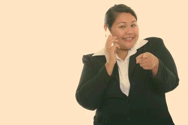 Studio plan de jeune heureuse grosse femme d'affaires asiatique souriant tout en parlant sur téléphone mobile et pointant vers la caméra — Photo