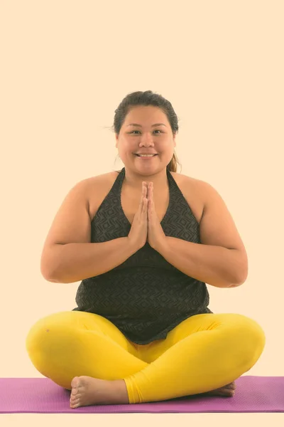 Studio shot de joven feliz mujer asiática gorda sonriendo y sentado mientras medita con ambas manos juntas —  Fotos de Stock