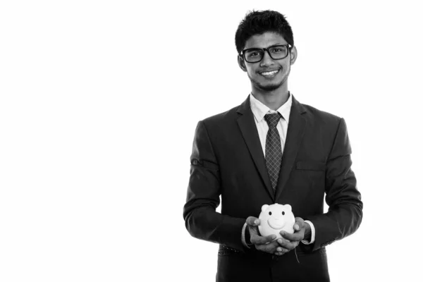 Studio shot of young happy Indian businessman smiling while holding piggy bank — Stock Photo, Image