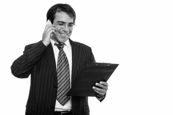 Młody szczęśliwy perski biznesmen uśmiecha się i rozmawia przez telefon komórkowy podczas czytania na schowku — Zdjęcie stockowe