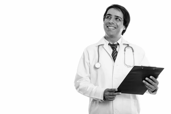 Estúdio tiro de jovem feliz persa homem médico sorrindo enquanto segurando prancheta e pensando — Fotografia de Stock