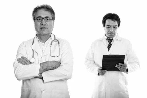 Studio shot of senior Persian man doctor with arms cross and young Persian man doctor reading on clipboard — Photo