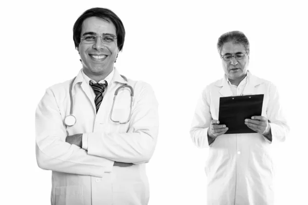 Joven feliz hombre persa médico sonriendo con los brazos cruzados y el hombre persa senior médico leyendo en el portapapeles —  Fotos de Stock