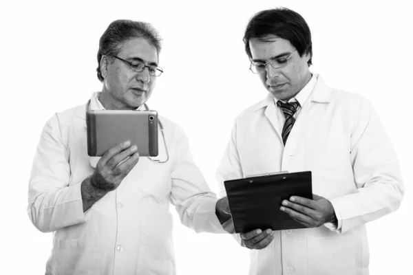 Studio shot of young and senior Persian man doctor holding digital tablet while reading on clipboard Royalty Free Stock Images