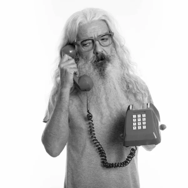 Studio shot of senior bearded man looking tired while talking on old telephone — Stock Photo, Image