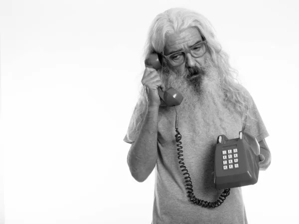 Studio shot of senior bearded man thinking and looking down while talking on old telephone — Stock Photo, Image