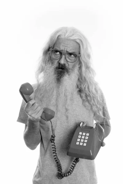 Studio shot of senior bearded man holding old telephone while thinking and looking up — Stock Photo, Image