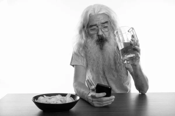 Studio shot of senior bearded man using mobile phone while holding glass of beer with bowl of potato chips on wooden table — Stock Photo, Image