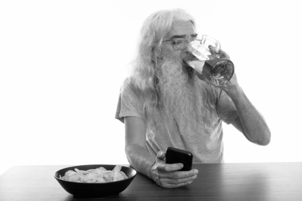 Студійний знімок старшого бородатого чоловіка, який використовує мобільний телефон, п'ючи келих пива з мискою картопляних чіпсів на дерев'яному столі — стокове фото