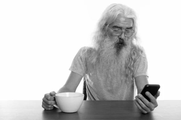 Estudio de tiro de hombre barbudo senior utilizando el teléfono móvil y la celebración de la taza de café en la mesa de madera — Foto de Stock