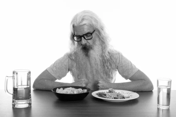 Estudio de tiro de hombre barbudo senior con anteojos mientras mira un tazón de papas fritas con alimentos saludables y poco saludables en la mesa de madera —  Fotos de Stock