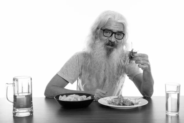 Studio shot di felice uomo barbuto anziano sorridente mentre mangia insalata con cibi sani e malsani sul tavolo di legno — Foto Stock