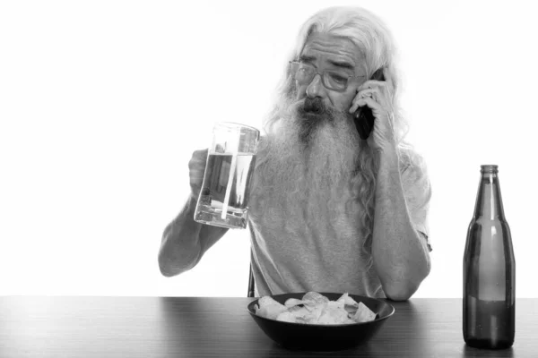 Studio shot of senior bearded man talking on mobile phone while holding glass of beer with bowl of potato chips on wooden table — Stock Photo, Image