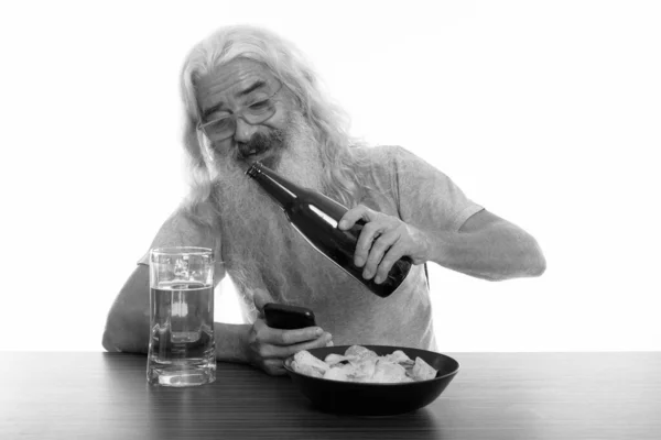 Photo studio d'un homme barbu heureux souriant tout en tenant son téléphone portable et en versant de la bière avec un bol de croustilles sur une table en bois — Photo