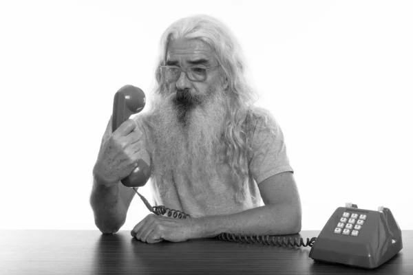 Studio shot of senior bearded man holding and looking at old telephone on wooden table — Stock Photo, Image