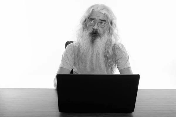 Estúdio tiro de homem barbudo sênior pensando ao usar laptop na mesa de madeira — Fotografia de Stock