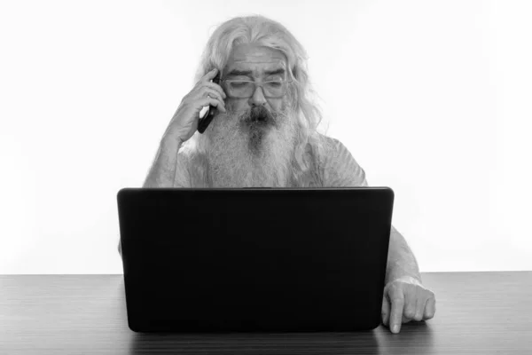 Studio shot van senior bebaarde man praten op mobiele telefoon tijdens het gebruik van laptop op houten tafel — Stockfoto
