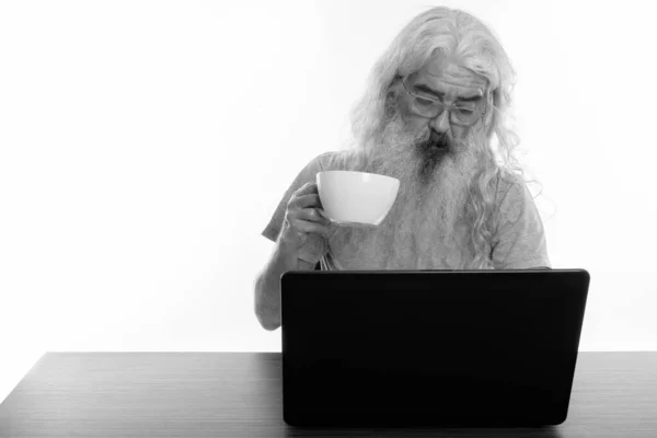 Plan studio d'un homme barbu âgé portant des lunettes et tenant une tasse de café tout en utilisant un ordinateur portable sur une table en bois — Photo
