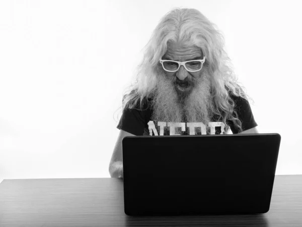 Plan studio d'un homme barbu âgé portant des lunettes et ayant l'air choqué en utilisant un ordinateur portable sur une table en bois — Photo