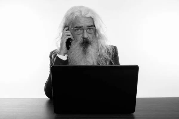 Plan studio d'un homme d'affaires barbu parlant sur un téléphone portable avec un ordinateur portable sur une table en bois — Photo