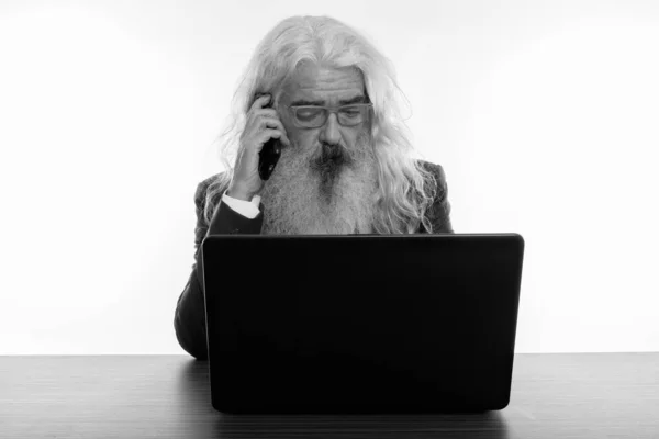 Plan studio d'un homme d'affaires barbu âgé parlant sur un téléphone portable tout en utilisant un ordinateur portable sur une table en bois — Photo
