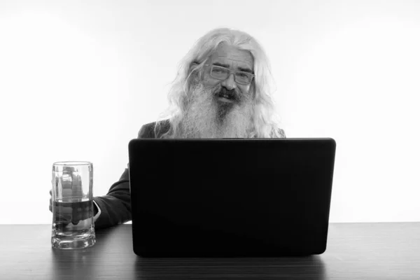 Plan studio d'un homme d'affaires barbu heureux souriant tout en utilisant un ordinateur portable avec un verre de bière sur une table en bois — Photo