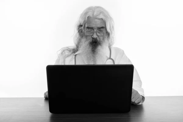 Studio zdjęcie starszego brodatego lekarza noszącego okulary podczas używania laptopa na drewnianym stole — Zdjęcie stockowe