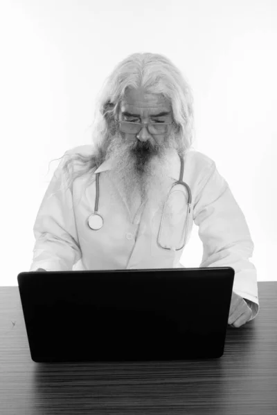 Studio shot of senior bearded man doctor using laptop on wooden table — Stock Photo, Image