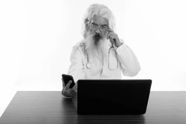 Yaşlı sakallı adamın cep telefonuyla ahşap masada dizüstü bilgisayar kullanırken çekilmiş bir fotoğrafı. — Stok fotoğraf