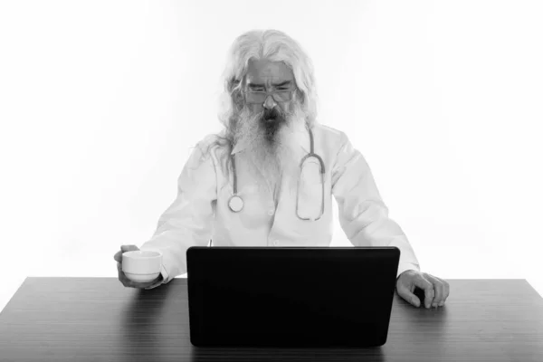 Studio shot of senior bearded man doctor holding coffee cup while looking at laptop on wooden table — Stock Photo, Image