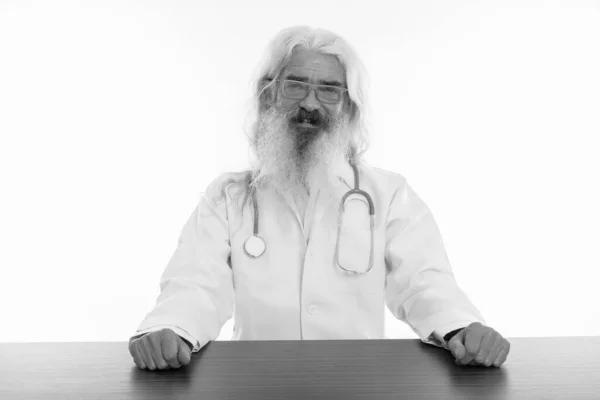 Studio colpo di felice anziano barbuto uomo medico sorridente mentre indossa occhiali e seduto sul tavolo di legno — Foto Stock