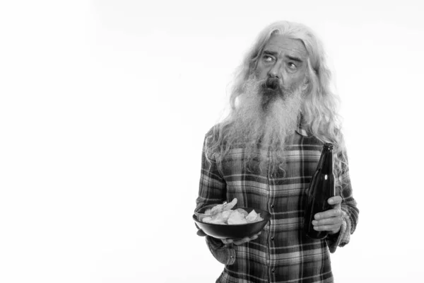 Studio shot di anziano uomo barbuto pensando, mentre in possesso di una bottiglia di birra e ciotola di patatine fritte — Foto Stock