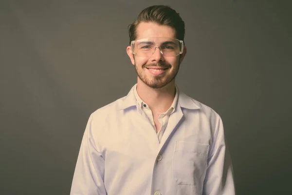 Joven hombre barbudo guapo médico con gafas protectoras contra el fondo gris —  Fotos de Stock