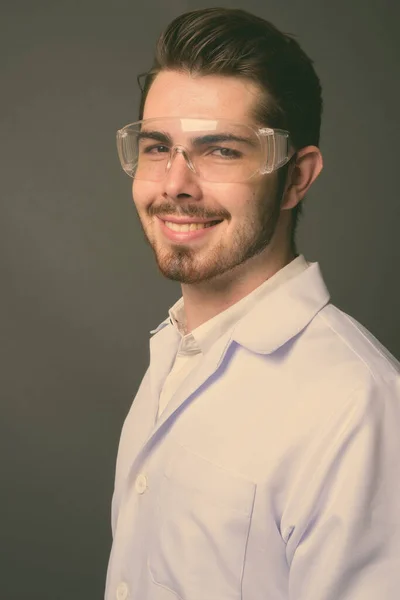 Giovane bello barbuto uomo medico indossa occhiali protettivi contro sfondo grigio — Foto Stock