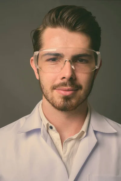 Mladý pohledný vousatý muž lékař nosí ochranné brýle proti šedému pozadí — Stock fotografie