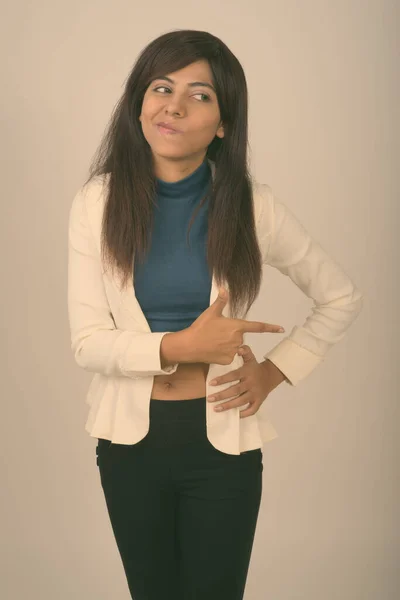 Studio shot of young Persian businesswoman standing and thinking while pointing to the side against gray background — Stock Photo, Image