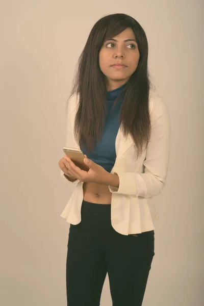 Studio shot of young Persian businesswoman thinking while holding mobile phone against gray background — Stock Photo, Image