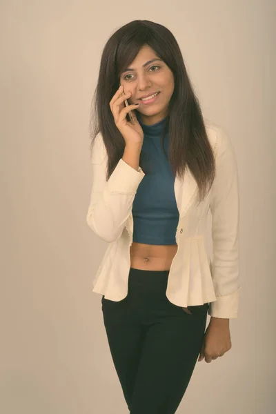 Studio colpo di riflessivo giovane felice donna d'affari persiana sorridente e in piedi mentre si parla sul telefono cellulare su sfondo grigio — Foto Stock