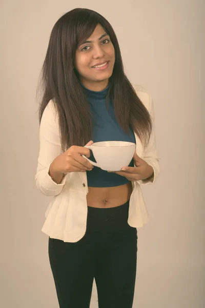 Studio shot di giovane donna d'affari persiana felice sorridente e in piedi mentre tiene in mano la tazza di caffè sullo sfondo grigio — Foto Stock