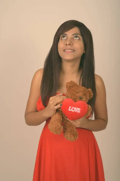 Young happy Persian woman smiling and thinking while holding teddy bear ready for Valentines day