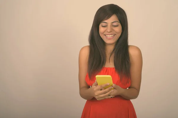 Studio colpo di giovane donna persiana felice sorridente durante l'utilizzo del telefono cellulare e indossa abito rosso sullo sfondo grigio — Foto Stock