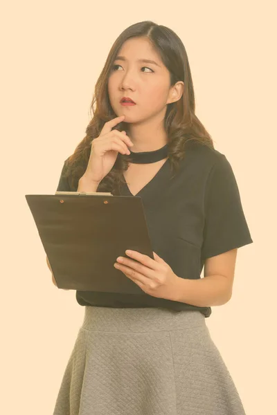 Joven hermosa mujer de negocios asiática sosteniendo portapapeles mientras piensa —  Fotos de Stock